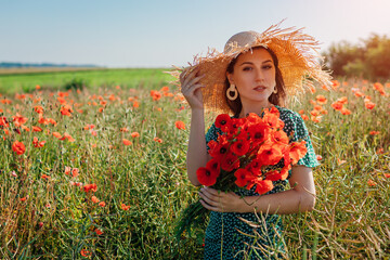 Stylish woman holding bouquet of poppies flowers walking in summer field. Fashionable girl wearing straw hat