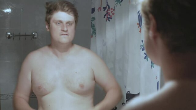 Young fat Caucasian man dances in front of the mirror and admires his reflection