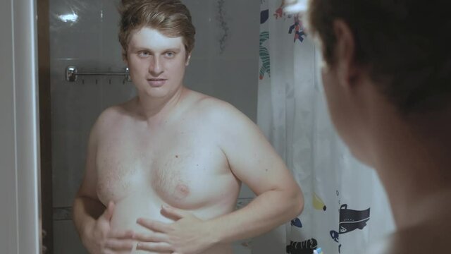 Young fat Caucasian man admires himself in the mirror and looks at his straining biceps