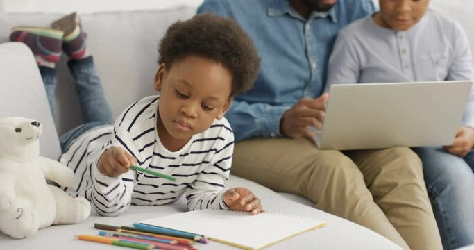 Cute African American little girl lying on couch and drawing picture with colorful pencils. Father and brother sitting beside and playing on laptop computer. Dad spending day with kids.