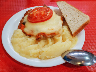 A plate with a baked piece of meat under cheese and tomato, with mashed potatoes and a piece of bread