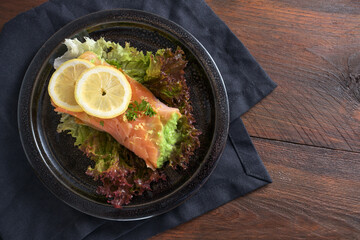 Low carb or ketogenic diet, smoked salmon filled with pea puree on lettuce salad with lemon slices and parsley garnish on a dark plate and napkin, rustic wooden table, copy space, high angle view