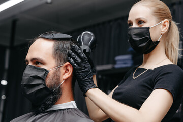 Woman barber cutting hair to a bearded man in face mask. Quarantine haircut concept.