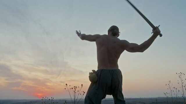 Muscular shirtless man with sword practicing slashing movements in a low angle view against the sunset sky in a side on low angle rear view