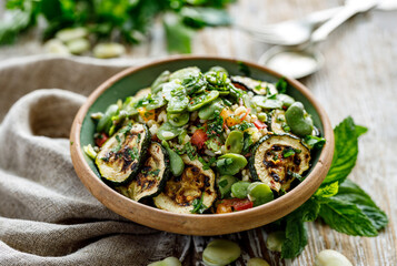 Broad bean salad with the addition of various vegetables sprinkled with fresh herbs in a ceramic bowl on a  wooden table close up view