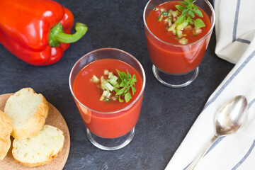 Summer diet. Cold tomato gazpacho soup in glass glasses with sliced cucumbers, peppers and greens on a black background top view. Sliced bread for snacks. Ice for cooling. Vegetarian food