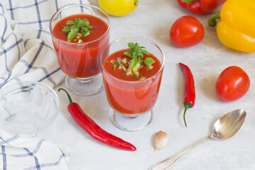 Summer cold tomato gazpacho soup in glass glasses with chopped cucumbers, peppers, lemon and greens on a white background. Ice for cooling. Vegetarian food