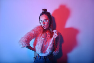 A girl in neon is dancing against a red-blue background. Hipster girl with fashionable haircut and glasses dancing, posing. Model in a fur coat at the club.