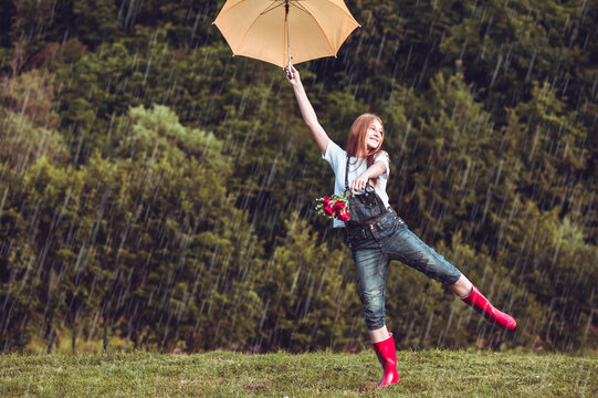 Happy ginger, teen girl with yellow umbrella and in red boots dancing the rain.