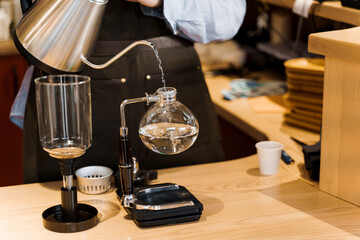 Fototapeta na wymiar Close-up barista pours hot boiling water from kattle in glass syphon device for coffee brewing in cafe. Syphon alternative method of making coffee. Scandinavian method of coffee making.