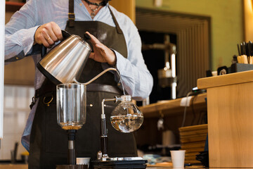 Close-up barista pours hot boiling water from kattle in glass syphon device for coffee brewing in cafe. Syphon alternative method of making coffee. Scandinavian method of coffee making.
