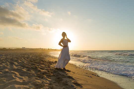 Backlit woman feeling free and happy, breathing clean fresh ocean air on a sandy beach in summer at dusk.