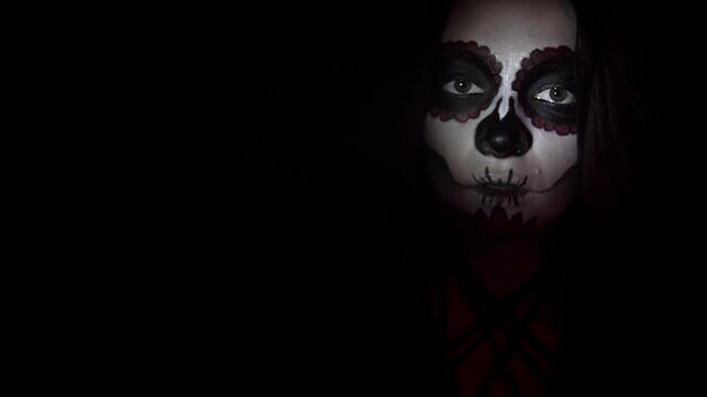Woman with makeup for the day of the dead smiles enigmatically in spotlight. Theme of costumes and transformation for holiday of Halloween. Traditional skull makeup for the day of the dead celebration