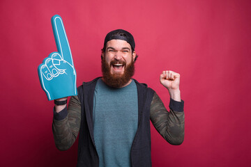 Happy man is making the winner gesture with a foam fan glove over red pink background
