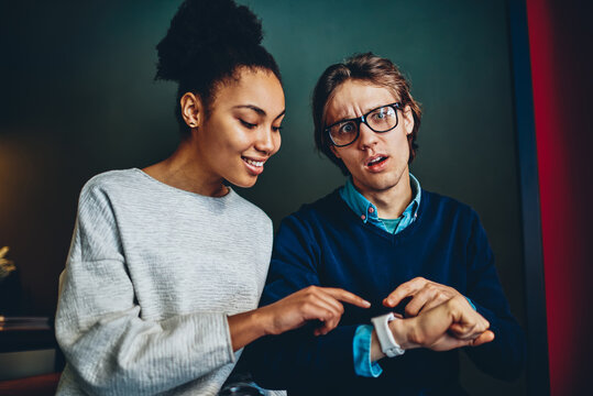 Young multiracial couple spending time together satisfied with advanced wearable computer, smiling woman pointing on smartwatch of stressed man shocked with notification from social networks. - Powered by Adobe