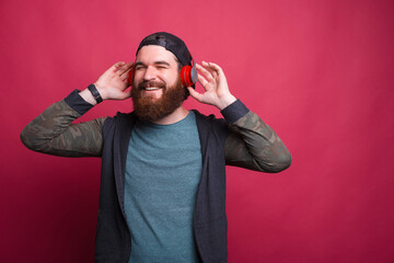 Happy smiling man is enjoying listening to the music in headphones with eyes closed over red background.