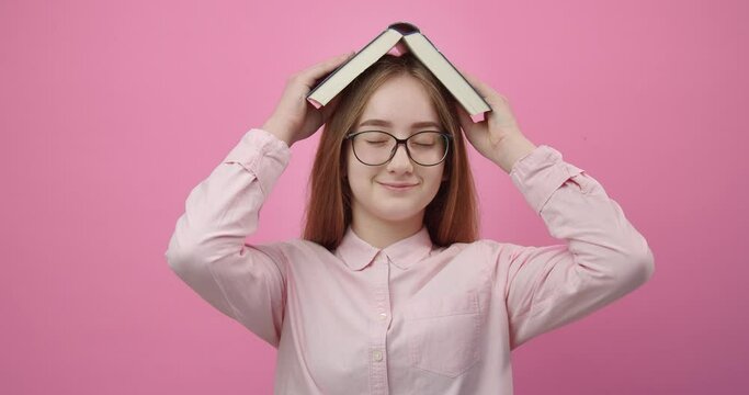Cheerful blonde student in pink shirt and eyewear holding opened book on head over pink background. Young girl having playful mood while posing in studio