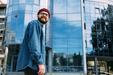 Happy young trendy bearded hipster male dressed in casual denim shirt, red hat and eyeglasses looking away and smiling