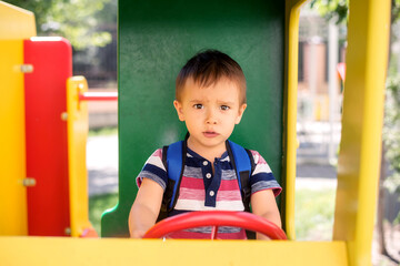 Fototapeta na wymiar Future driver: toddler boy with serious face playing as truck driver at outdoor playground. Little kid with backpack sitting at steering wheel and driving toy vehicle in park. Child dreams concept