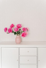 big bouquet of fuchsia peonies in a pink vase in a bright room are on a white chest of drawers. cozy house, fresh flowers at home, home greenhouse, minimal design, loft house