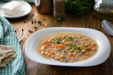 Vegetarian thick cheakpea soup with corrots and greens in white plate on wooden table with ingredients around. Turkish and greek cuisine. Healthy vegetable food. Horizontal. Etli Nohut, Revithia dish.