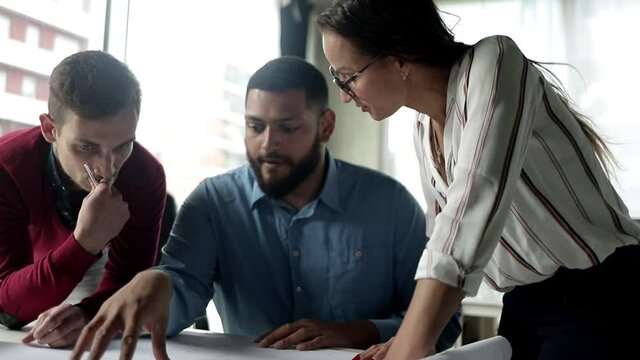 Serious construction expert explaining blueprint details to colleagues. Business people standing at table and discussing building floor plan. Medium shot. Engineering or teamwork concept