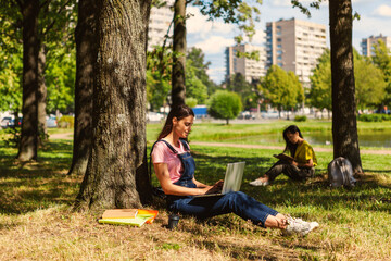 Fototapeta premium Smiling college girl sitting under tree leaning on trunk and studying on laptop in park. Another female student reading book in background