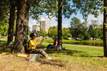 Side view of female student in VR headset swiping visualized data sitting under tree in park. Another college girl wearing virtual reality glasses in background