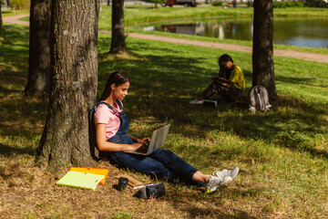 Naklejka na ściany i meble Side view of female student sitting under tree and typing on laptop in parkб textbooks, coffee cup and VR glasses nearby. Another college girl reading book in background