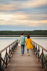 Rear perspective view of young romantic couple walking on wooden pier over river holding hands in...
