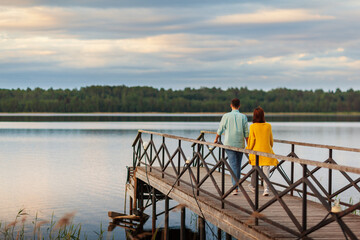 Back view of loving young couple walking on old wooden pier over river holding hands in summer evening