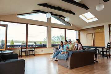 Group of four young friends sitting on sofa in countryside house, eating tasty pizza, watching tv show or series and talking, side view wide shot