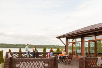 Two young couples relaxing on wooden country house terrace. Couple talking sitting at table, their friends talking over drink leaning on railing in background. Wide shot with copy space on sky