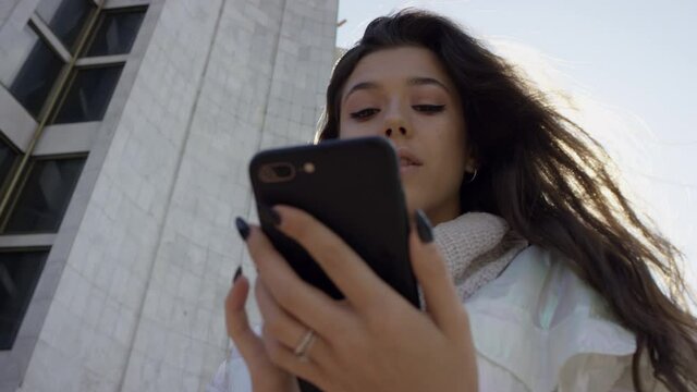 A young dark-haired woman standing near the entrance of a building abd sending an e-mail to her friend on the phone. A bottom view slow motion close up shot