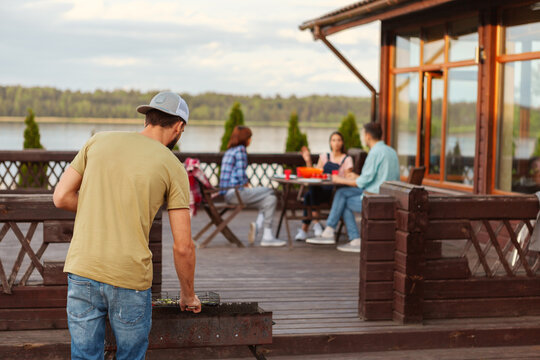 Back View Of Young Man Cooking Grilled Vegetables. Group Of His Friends Sitting At Table On Wooden Terrace Of Country House And Talking At Bbq Party