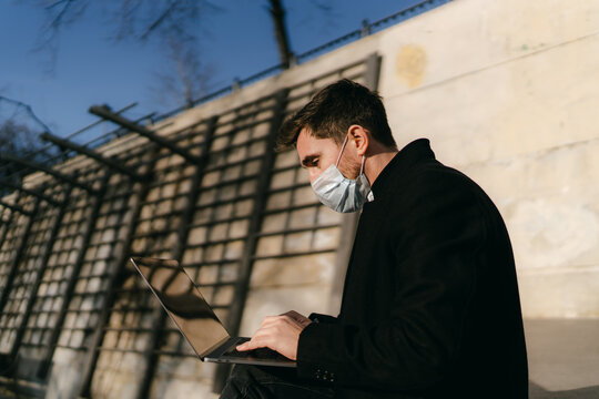 A man sits on the street wearing a medical mask and works behind a laptop during coronavirus quarantine