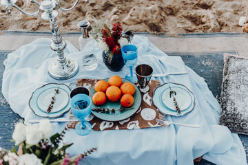 Maritime wedding picnic with decor, candles, flowers, vases, oranges, lavender, red wine in blue glas on the carpet at beach.