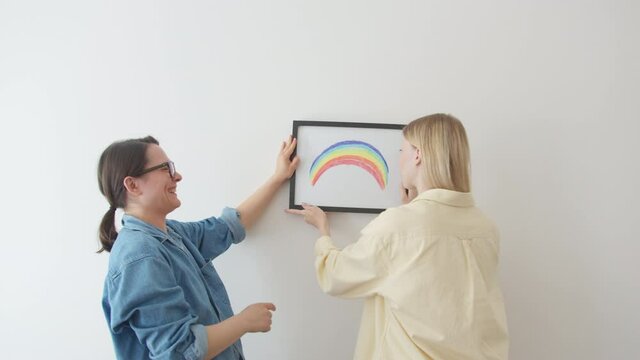 Close up Cute and happy young couple lesbian, during home remodeling or moving new apartment, hangs picture Rainbow flag on wall. Enjoying team work together. Girls are happy together
