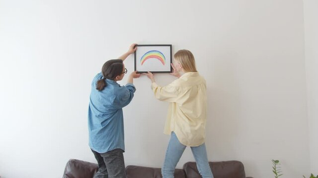 Cute beautiful and young couple lesbian, during home remodeling or moving new apartment, hangs picture Rainbow flag on wall. Enjoying team work together