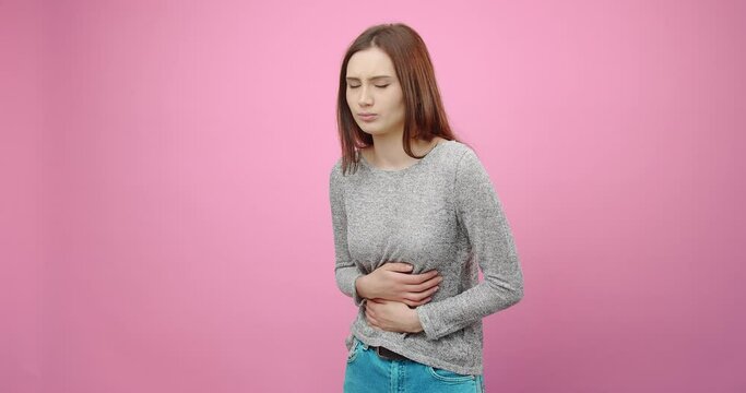 Dissatisfied young girl in casual outfit suffering from stomach ache while standing over pink background. Upset woman with dark hair feeling discomfort in studio.