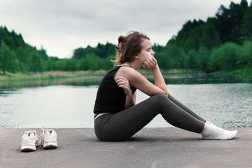 Teenage girl in a bad mood sitting on pond pier in cloudy summer morning. The girl is worried about something. Awkward age.