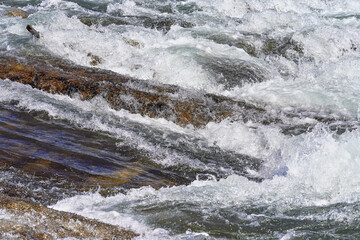 Rapid spring river flowing over rocks on sunny day, forming white water waves, closeup detail - abstract nature background