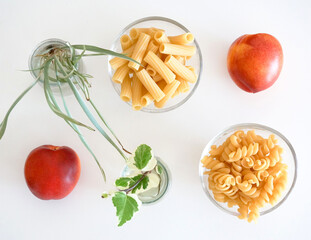 Raw pasta in tiny glass bowls, sweet nectarins and flower offsets plants on white background. Food...