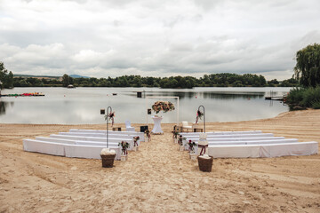 Wedding ceremony at lake - white benches and flower decoration on sandy beach. Outdoor nuptials. Wedding day concept.