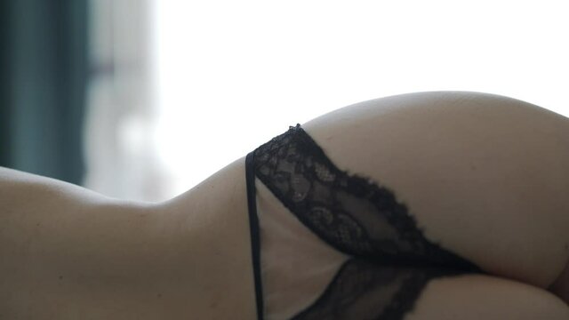 Rear view of attractive young caucasian woman in sexy lingerie lying on bed. Beautiful female body in lace black panties close up. Female bare back against window with curtain moved by the wind.