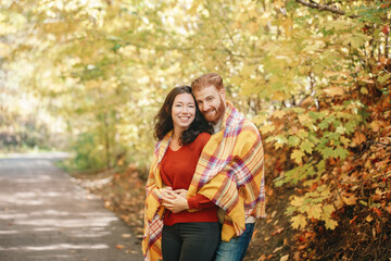 Fototapeta premium Beautiful couple man woman in love. Boyfriend and girlfriend wrapped in yellow blanket plaid hugging together in a park on autumn fall day. Tenderness and happiness. Authentic real people.