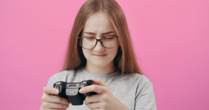Portrait of excited young girl in eyeglasses using joystick for playing video games. Isolated over pink studio background. Concept of addiction and entertainment.