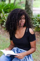 a young black woman between 20 and 30 years old sitting reading a book alone, in a park