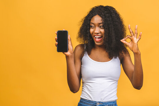 Portrait of a smiling young african american black woman holding blank screen mobile phone and showing ok sign isolated over yellow background.