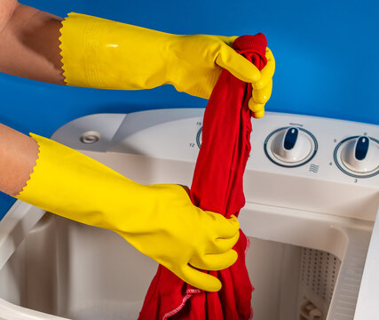hands with yellow gloves squeezing a red shirt from a white washing machine and a blue background